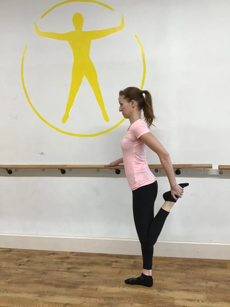 Woman standing upright , holding onto a bar with her left leg bent and holding onto her foot
